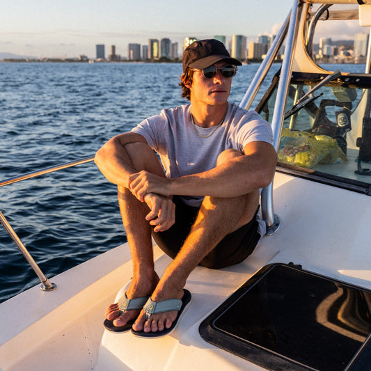 Man wearing seagreen and trench blue swim shorts sitting on a boat deck with ocean and cityscape in background.