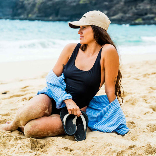Woman wearing ecru baseball cap and black sleeveless swimsuit top on sandy beach with ocean in background.