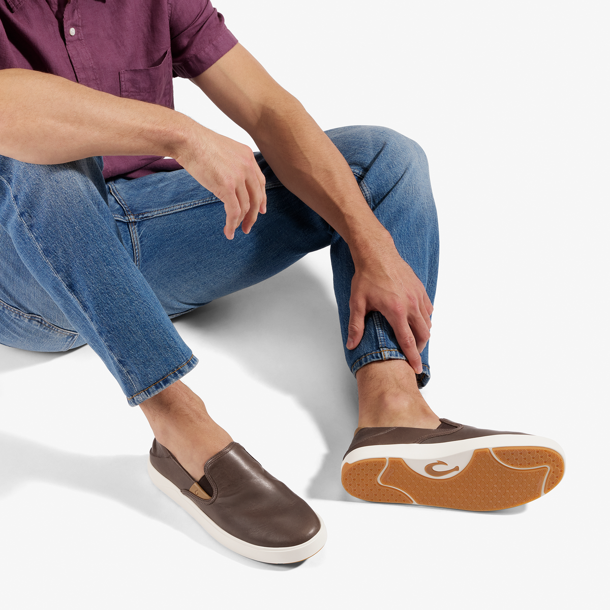 Person wearing dark brown slip-on sneaker with white sole, blue jeans, and burgundy shirt, shown from waist down.