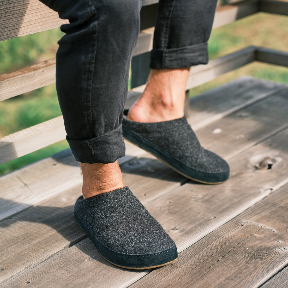Person wearing dark gray wool slippers with dark jeans cuffed at the ankles, standing on wooden deck.