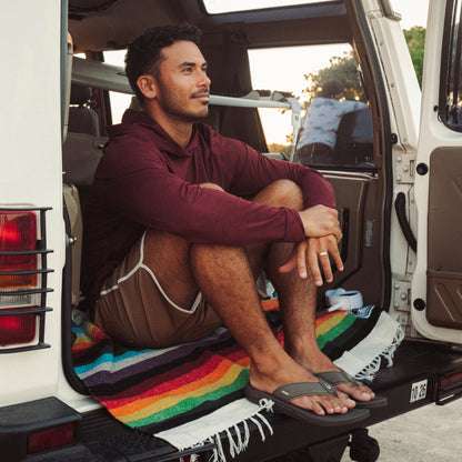 Man in maroon hoodie and brown shorts sitting in open van door wearing brown koa sandals.