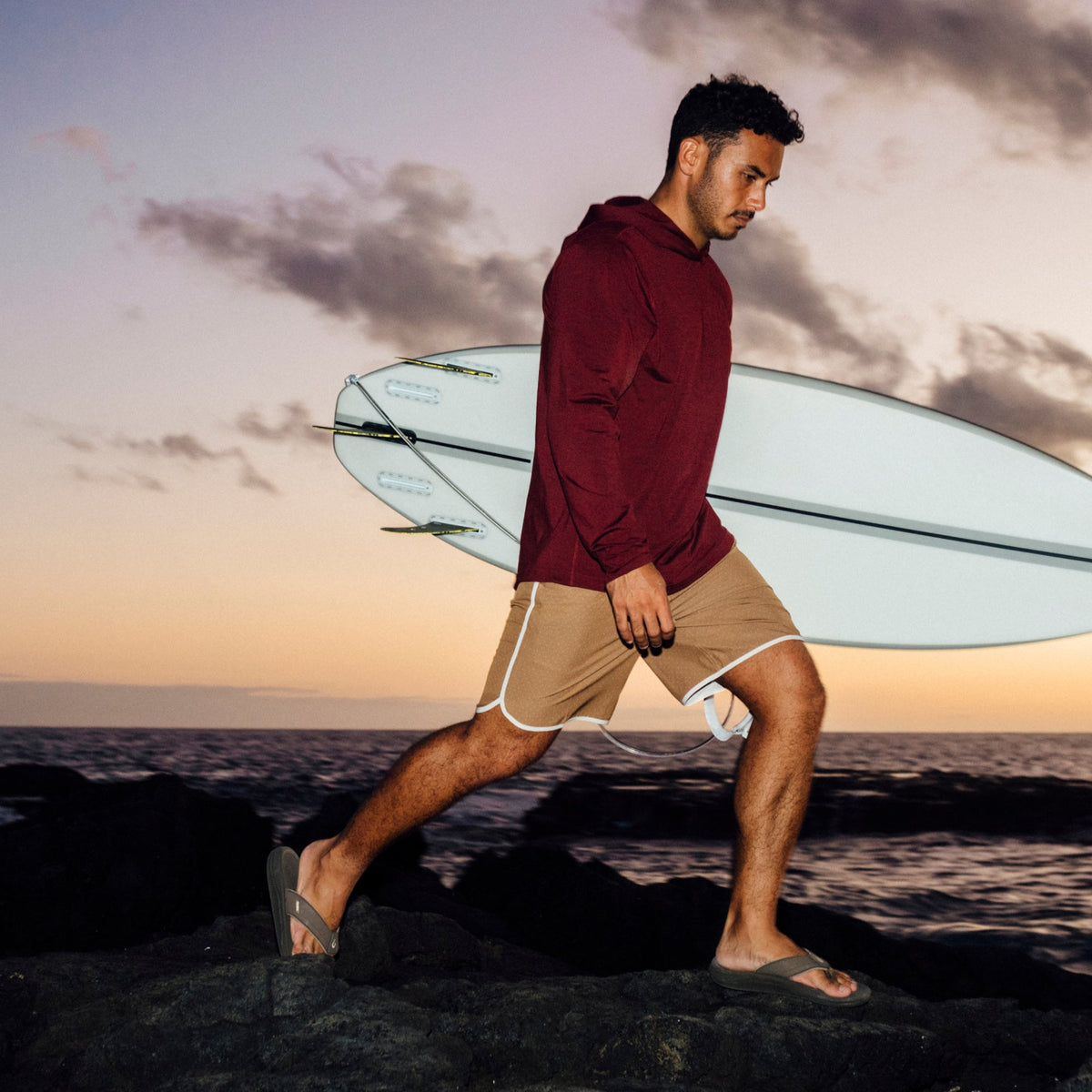 Man in maroon hoodie and tan shorts carrying brown koa surfboard on beach at sunset.