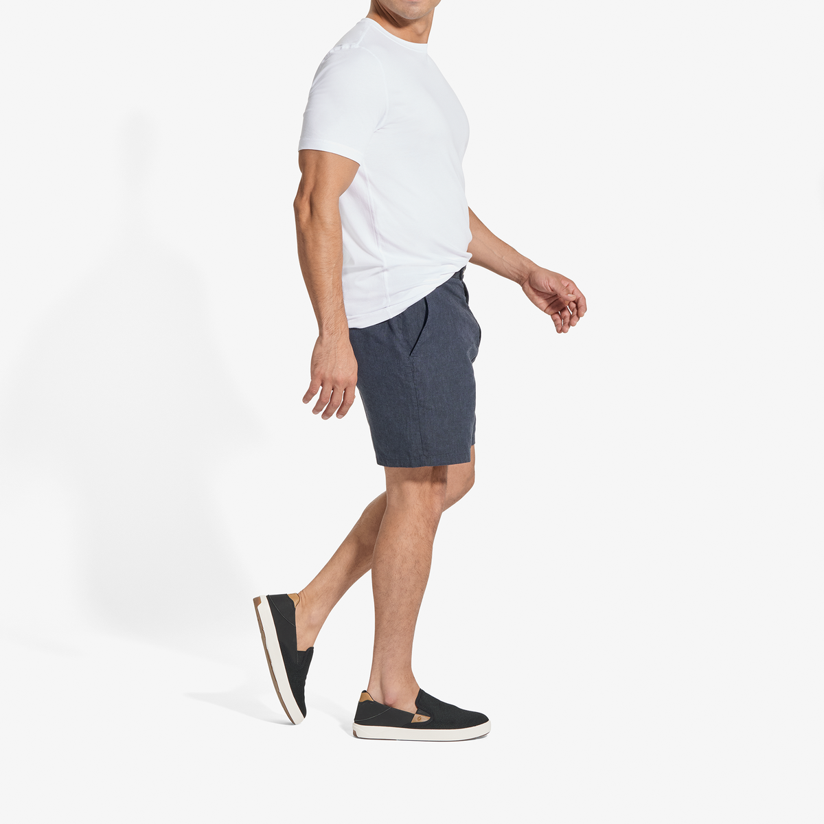 Man wearing white short-sleeve t-shirt, dark blue shorts, and black slip-on sneakers with tan trim.